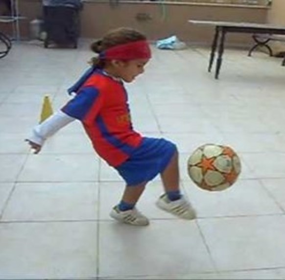 This photo shows a youngster kicking a soccer ball.  The child’s arms are held backward while the head is tipped forward.