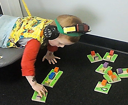 this photo shows a child who is prone on a platform swing.  He is playing a game with wooden car & truck dominoes.  The vehicles are posed in different spatial orientations.
