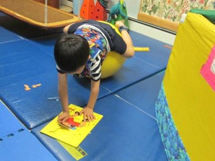 this photo shows a boy who is lying on his stomach over a physioball. His back and neck are extended while he engages the spatial orientation of puzzle pieces.