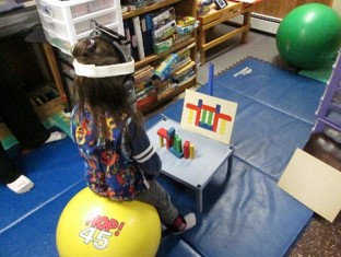 this photo shows a child who is sitting on a physioball while engaging spatial orientation of building a tower of blocks.  The back is extended while the head is straight upright.