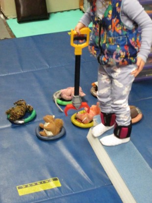 this photo shows a child who is standing erect on a balance beam while using a reacher to place small stuffed animals into rings.