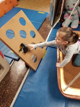 his photo shows a girl who is kneeling on a platform swing, reaching toward a target board of holes to push a stuffed animal through one of the holes.