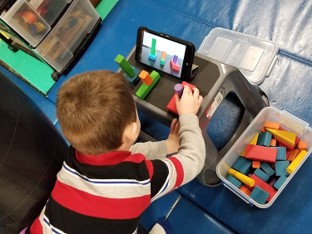  this photo shows a boy who is reaching to build a tower of blocks.  He is sitting on a tire swing and his back and head are extended as he reaches to assemble the blocks as shown on a tablet.