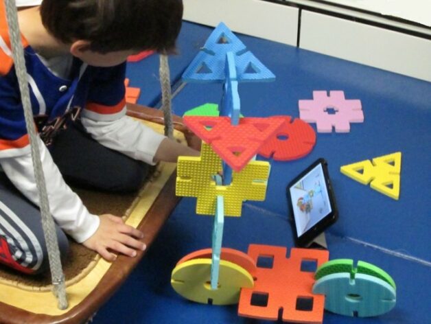 This is an image of a child who is using large foam shapes to build a toy scooter that matches the steps shown on a tablet