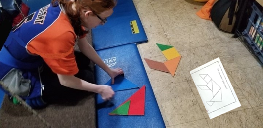This image shows a girl at work on the floor .  She is using large felt tangram pieces to  construct a puzzle of a polar bear from an outline.