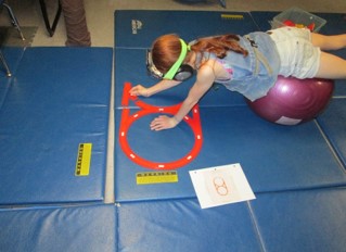 This image of a girl who is prone lying over a physioball is strengthening her back while copying track layouts from a diagram