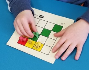 The photo shows one hand holding a task card still while the other hand picks up and places a small toy on the task card