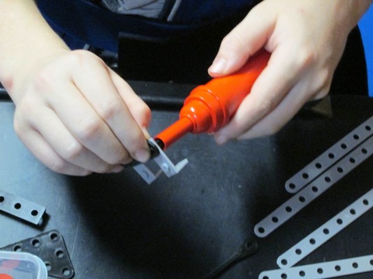 This photo shows use of a toy rivet gun to connect two strips of a building project together.  One hand is holding the two strips of the project still, while the other hand is manipulating the rivet gun.  