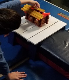 This is a photo of a boy who is holding his body steady with one hand while the other hand is being used to position a Lincoln Log on top of a log cabin.