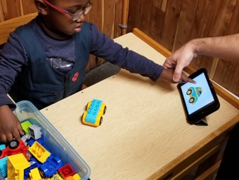 This photo shows a boy who is pointing to an image on the iPad as he selects corresponding Duplo blocks to build a copy of the  car shown on the iPad
