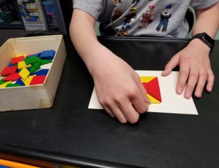 the  photo shows a boy using both hands to arrange parts of a parquetry puzzle.  One hand is holding the task card still while the other hand moves the pieces