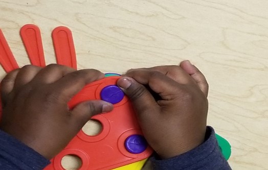 This photo shows how a young  child’s hands learn to push a button through a button hole while using the Busy Buttons toy.  One hand is holding the hole still while the other hand is pushing the button through the hole.