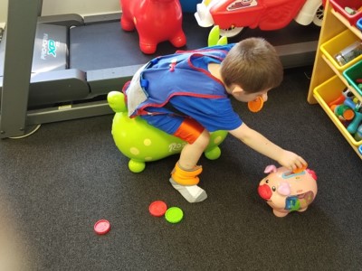 This picture shows a boy who is leaning over from a hop horse riding toy to pick up tokens and put them into a piggy bank