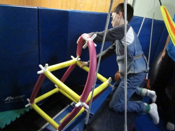 This picture shows a boy who is on a swing and reaching over to add a long pool noodle to a rocket ship he is building