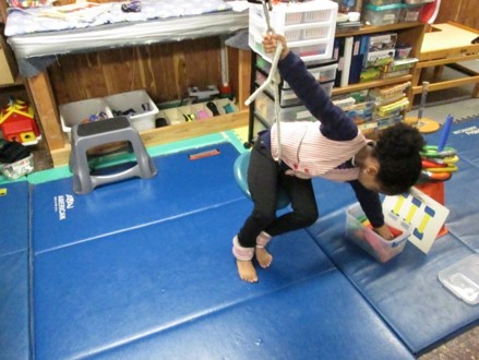 This picture shows a girl leaning over to pick up a block while she is sitting on a disk swing.    One holding onto the suspended rope while the other hand is picking up the block she needs to complete the and is project