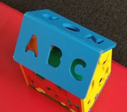 this is a photo of an Alphabet House. It is a toy with  cut out shapes for letters of the alphabet.  It is the target for the girl who is kneeling in back of the exercise ball.