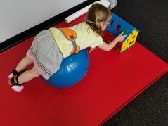 this photo shows the girl rolling onto her belly to place the letter “C” into the corresponding cut out located on the roof of the Alphabet house