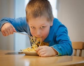 this photo shows a boy who is eating spaghetti with a fork.  He is shoveling the food into his mouth in a very messy way