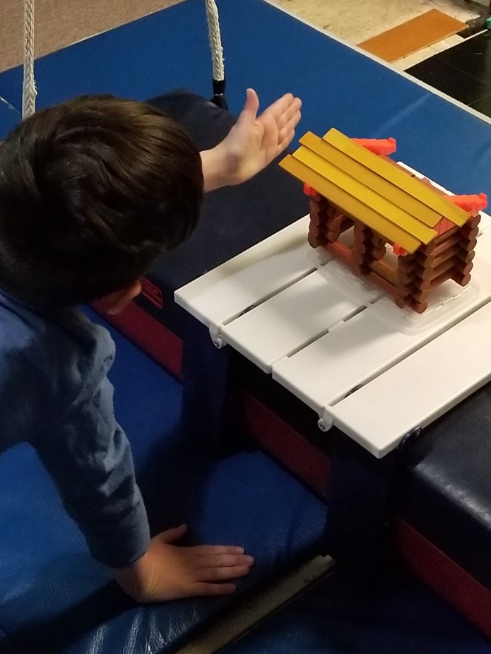 the photo of the boy shows his plan to tap the roof slats gently in order to align them as he completes the top of the Lincoln Log cabin he has built.