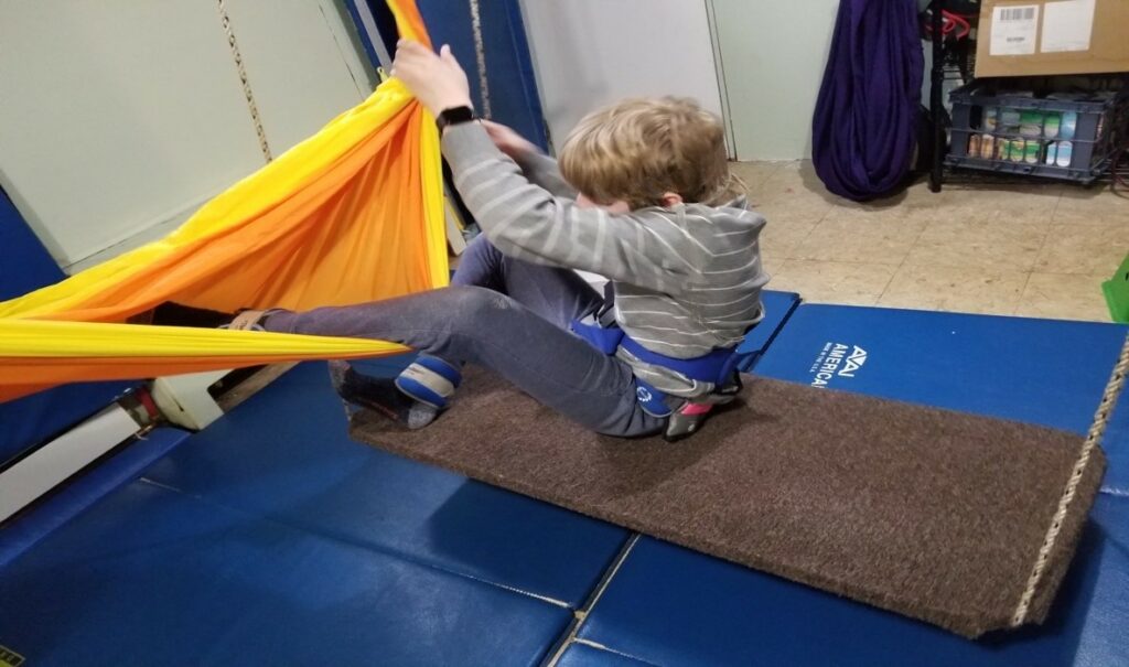 This photo shows a boy who will be moving from one swing into a hammock.  He is  sitting on a platform swing and using both arms to hold the  nearby  hammock open while he moves his legs into the center of the hammock in preparation for transitioning his whole body  into the center of the hammock. 