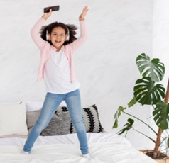 This photo shows a girl jumping on her bed while dancing with headphones.  Her arms are pushed up and out while sings along to add even more input.