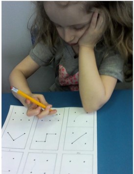 The picture shows a young girl trying to copy a diagram.  She is using one hand to hold her head up, and the other hand to grip the pencil to draw the diagram.  Her pencil grip is very awkward.