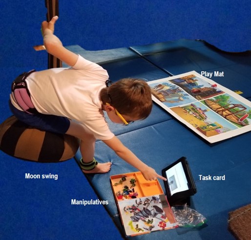 the photo shows a boy sitting on a teardrop shaped swing. He is  keeping  himself steady by holding onto the top of the swing with his left hand, while using his right hand to point to the task card .