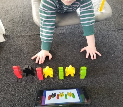 This photo a young boy using a walk out from a doorway swing while he is arranging a lineup of robots as shown on a  digital task card.
