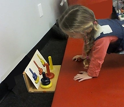 This photo shows a young girl using a  walk out to select and stack alternating color size  rings onto colored poles as shown by a task card.