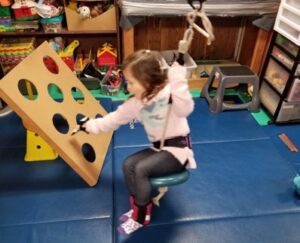 This young girl is holding the rope of a swing to pull the weight of her body up against gravity while she swings forward to toss a toy into a hole.