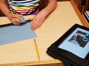 The photo shows a youngster tracing a line along the edge of a ruler that is positioned at the bottom of a piece of construction paper.  One hand is holding the paper still while the other hand is holding the pencil and drawing the line needed. 