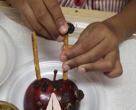 the photo shows a girl making a food craft.  She is using an apple that has been cut in half while a  pretzel has been pushed into the front as antenna for a ladybug. The photo shows her using one hand to hold a pretzel stick still with one hand, while using the other hand to push a blueberry on top of the bugs’  “antenna”