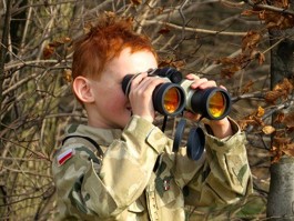 the photo shows a boy scout using visual fixation skills as he uses a pair of binoculars to look at the surrounding woodlands.