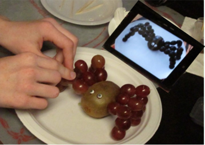 picture of a child using a kiwi and grapes to recreating a spider from an image in on an iPad.