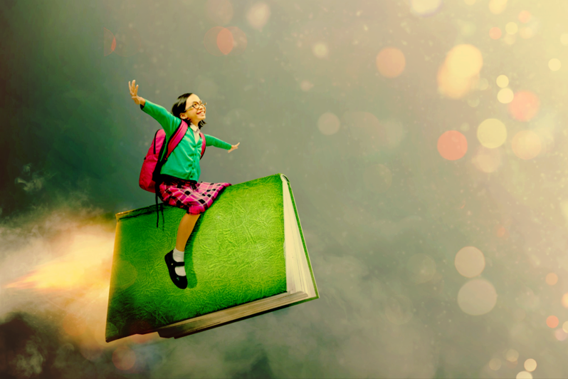 The image above shows a girl with a backpack sitting straddled upon a book with her arms stretched out against the background of a cloud filled sky